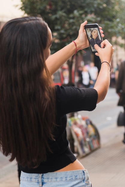 Vista traseira, de, um, mulher jovem, levando, selfie, ligado, telefone móvel, em, rua