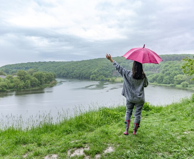 Vista traseira de uma garota sob um guarda-chuva em uma caminhada na floresta perto do lago em tempo chuvoso