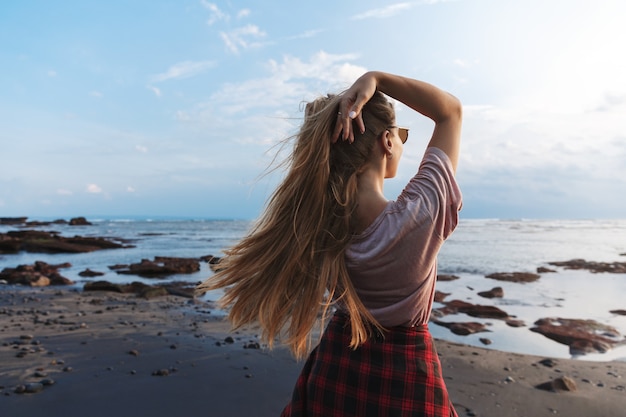 Foto grátis vista traseira de uma garota viajante com cabelo comprido, apreciando a vista do mar azul em pé de praia de areia vulcânica preta.