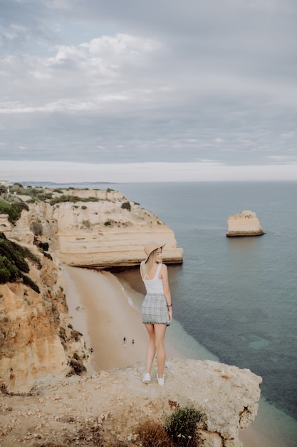 Vista traseira de uma jovem com um chapéu e um lindo vestido de verão em pé na pedra