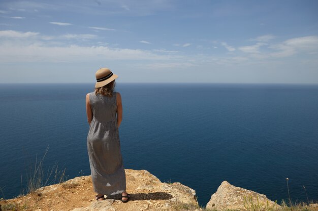 Vista traseira de uma linda mulher usando chapéu de palha e vestido maxi de verão em pé no topo da montanha e contemplando o mar infinito magnífico na frente dela. Férias, viagens e conceito à beira-mar