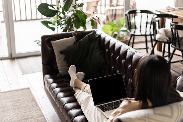 Vista traseira de uma mulher asiática sentada em casa e descansando na sala de estar, deitada no sofá-sofá com o laptop.