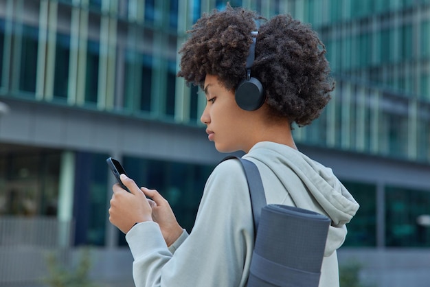 Vista traseira de uma mulher desportiva de cabelo encaracolado ouve música motivacional através de conversas online com auscultadores num smartphone vestido com roupa desportiva
