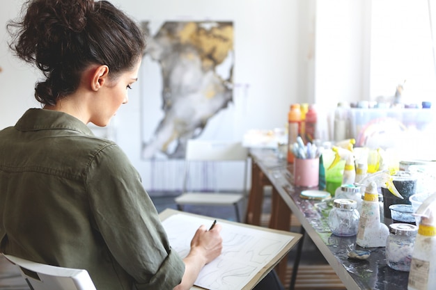 Foto grátis vista traseira do artista morena jovem mulher caucasiana em camisa cáqui, segurando o lápis, desenhando na oficina com tintas na mesa perto dela. conceito de arte, criatividade, pintura, hobby, emprego e ocupação