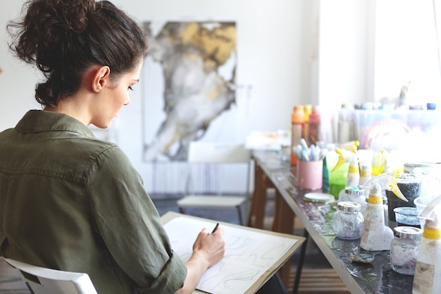 Vista traseira do artista morena jovem mulher caucasiana em camisa cáqui, segurando o lápis, desenhando na oficina com tintas na mesa perto dela. Conceito de arte, criatividade, pintura, hobby, emprego e ocupação