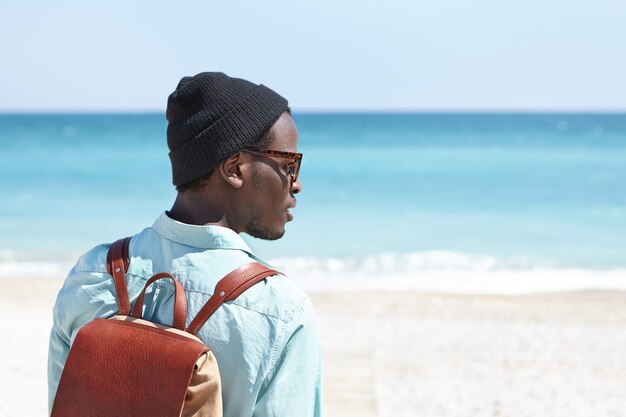 Vista traseira do despreocupado jovem viajante de pele escura com mochila de couro, desfrutando de uma bela vista do mar azul enquanto passa as férias de verão à beira-mar, contemplando uma vista incrível em dia ensolarado