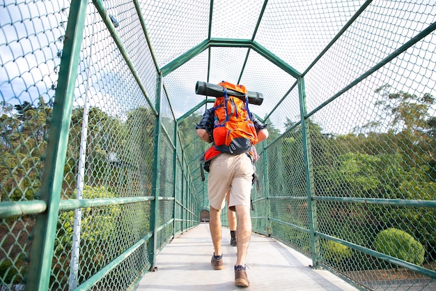 Vista traseira do homem andando na ponte rodeada por grade verde. Caminhantes carregando mochilas, atravessando rios ou lagos pelo caminho. Turismo de mochila, aventura e férias de verão