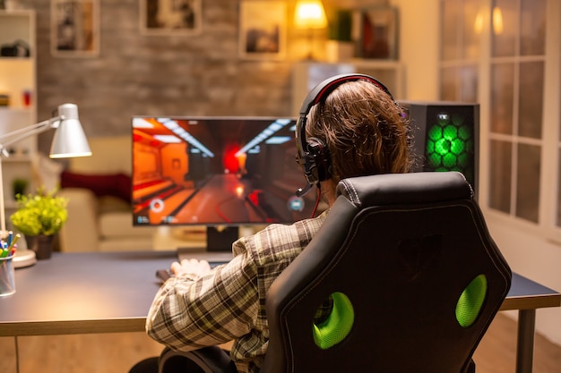 Vista traseira do jogador masculino jogando um atirador em seu poderoso computador PC tarde da noite na sala de estar.