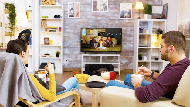 Vista traseira do jovem casal sentado em cadeiras confortáveis, comendo macarrão na frente da tv usando os pauzinhos. Gato dormindo em sua cama aconchegante.