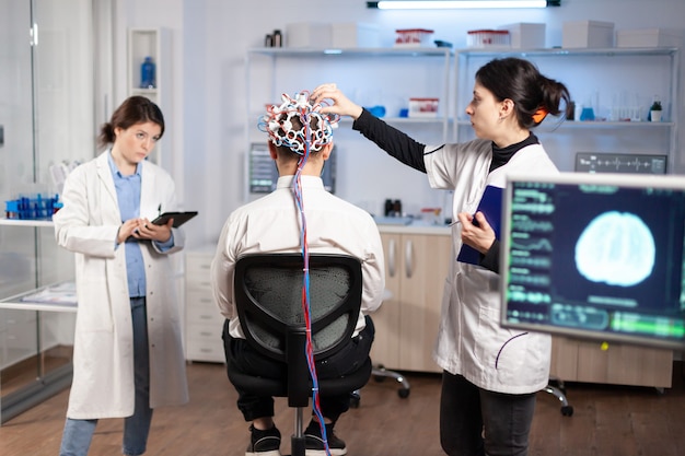 Vista traseira do paciente homem usando fone de ouvido de varredura de ondas cerebrais de desempenho sentado no laboratório de pesquisa neurológica enquanto o pesquisador médico o ajusta, examinando o sistema nervoso, digitando no tablet.