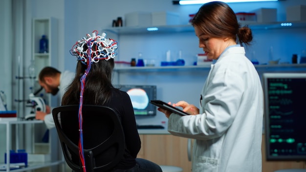 Vista traseira do paciente mulher usando fone de ouvido de desempenho eeg sentado na cadeira no laboratório de pesquisa neurológica enquanto o pesquisador médico o ajusta, examinando o sistema nervoso, digitando no tablet.