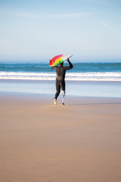 Vista traseira do surfista deficiente indo para o mar com a prancha. Homem ativo com uma perna amputada segurando uma prancha de surfe e surfando nas férias de verão