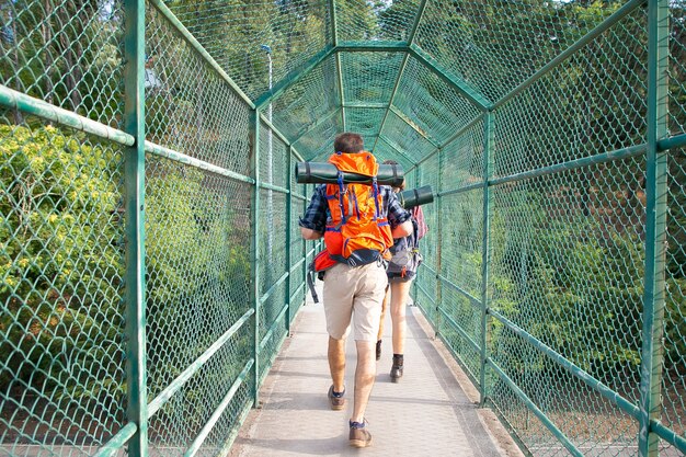 Vista traseira dos caminhantes andando na ponte cercada com grade verde. dois turistas carregando mochilas e passando por um caminho. conceito de turismo, aventura e férias de verão