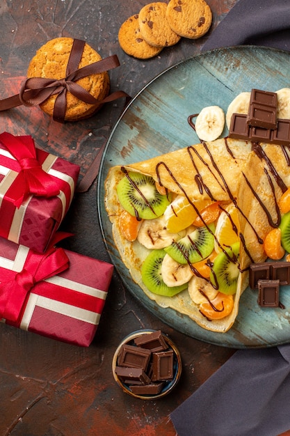 Vista vertical de um delicioso crepe servido com frutas cítricas picadas, decorado com calda de chocolate e caixas de presente em cores diferentes