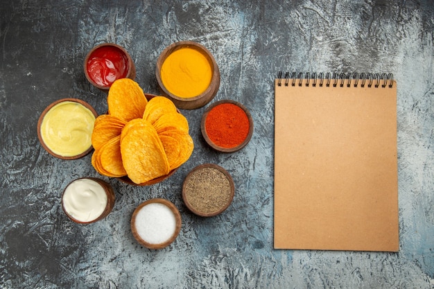 Visualização horizontal de chips crocantes na tábua de madeira servidos com ketchup de maionese de diferentes especiarias e caderno na mesa cinza