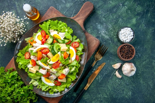 Visualização horizontal de uma deliciosa salada com muitos ingredientes frescos em uma tábua de corte de madeira, especiarias, frasco de óleo, talheres, em, fundo