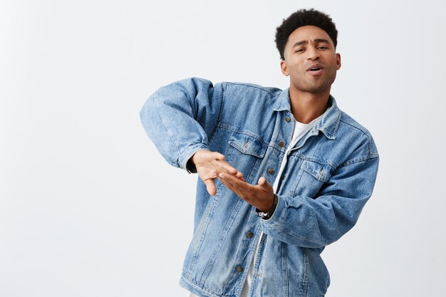 Vivendo uma vida rica. Emoções positivas. Retrato de jovem homem de pele negra atraente com penteado afro em jaqueta jeans e camiseta branca, jogando dinheiro fora para o videoclipe, se divertindo com os amigos