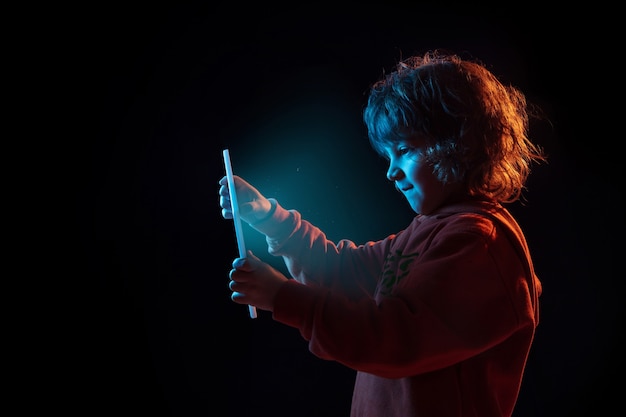 Vlogging com tablet, jogar. Retrato do menino caucasiano em fundo escuro do estúdio em luz de néon. Lindo modelo cacheado. Conceito de emoções humanas, expressão facial, vendas, anúncio, tecnologia moderna, gadgets.