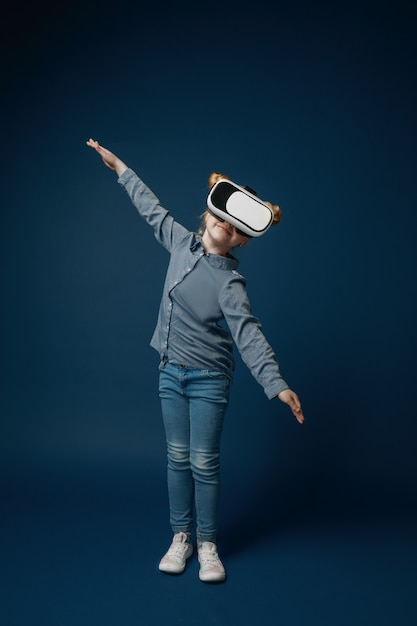 Voando como uma aeronave. Menina ou criança em jeans e camisa com óculos de fone de ouvido de realidade virtual, isolados no fundo azul do estúdio. Conceito de tecnologia de ponta, videogames, inovação.