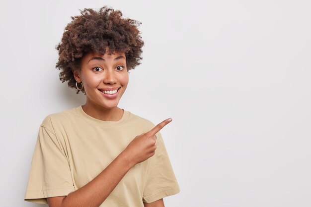 Você deveria dar uma olhada. Mulher bonita sorrindo alegremente aponta o dedo indicador no canto superior direito mostra a melhor escolha de produto promocional vestido com uma camiseta casual bege isolada sobre fundo branco