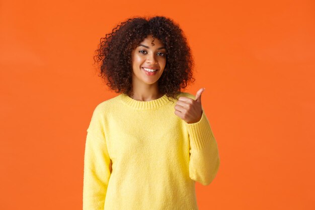 Você deveria vir e ver. Suéter amarelo despreocupado e bonito de menina hippie, convidando para entrar apontando o polegar atrás de si e sorrindo, recomendar um produto ou serviço da empresa, fundo laranja