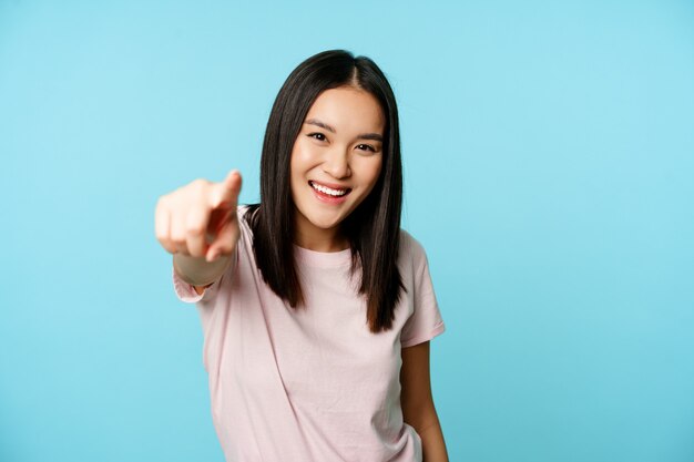 É você. Mulher asiática feliz sorridente, apontando o dedo para a câmera, parabenizando, convidando as pessoas, em pé na camiseta sobre fundo azul.