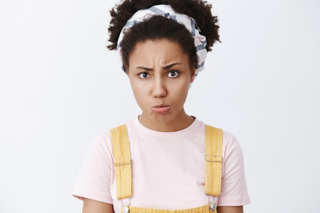 Você prometeu passar um tempo comigo. Retrato de uma mulher afro-americana triste e ofendida, com bandana e macacão amarelo, franzindo os lábios, carrancuda e amuada por causa da ofensa, sobre uma parede cinza