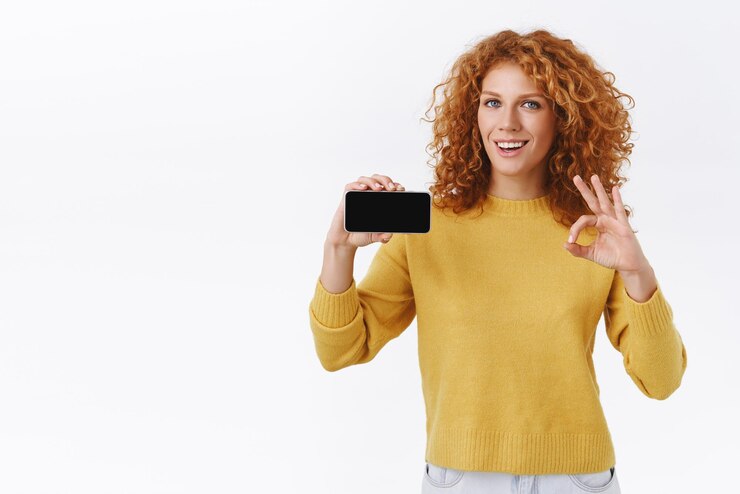 Foto grátis você tem que experimentá-lo atraente ambiciosa ruiva encaracolada de suéter amarelo segurando o smartphone horizontalmente mostrar sinal de ok ok e sorrindo em aprovação recomendo taxa de aplicativo bom perfil de jogo