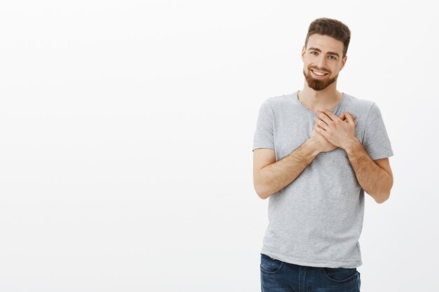 Você tocou meu coração. Retrato de bonito, elegante e carismático masculino caucasiano moreno com barba segurando os braços no peito agradecido e agradecido sorrindo amplamente com sincero amor e admiração