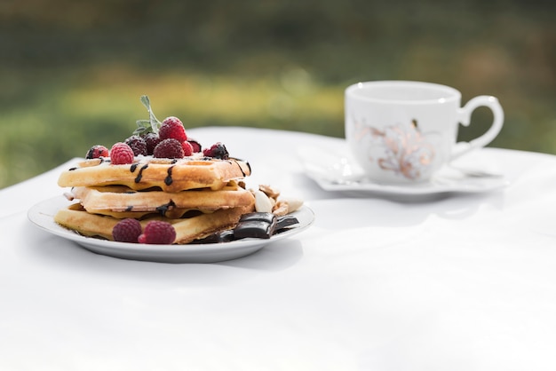 Waffles com coberturas de framboesas na placa e café de cerâmica na mesa branca no exterior