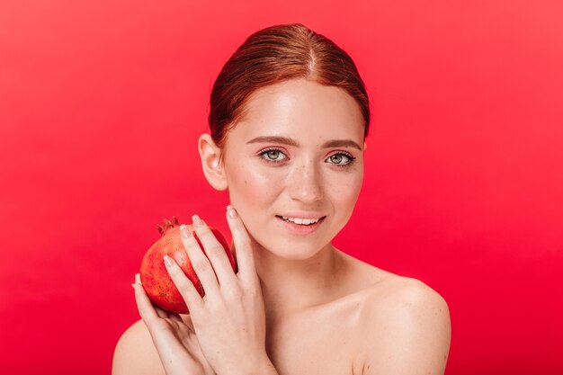 Winsome garota segurando granada com um sorriso gentil. Foto de estúdio da incrível senhora gengibre com frutas isoladas em fundo vermelho.