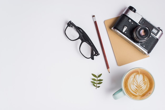 Xícara de café com leite; lápis; óculos e câmera vintage no fundo branco