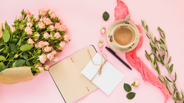 Xícara de café em cerâmica; buquê rosa; diário e cartão no pano de fundo rosa