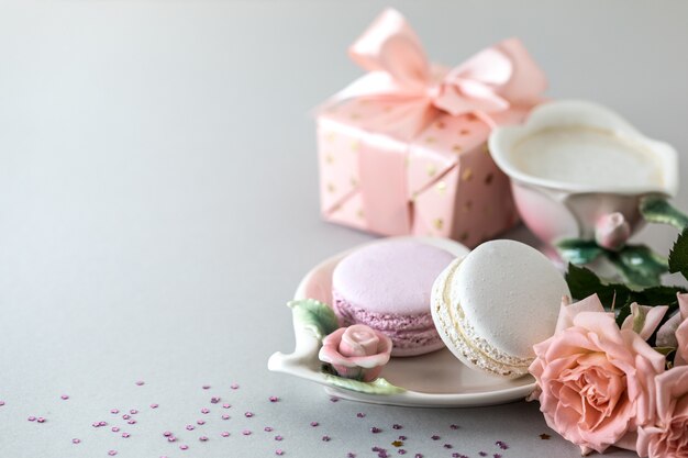 Xícara de café, macarrão para bolo, um presente em uma caixa e rosas cor de rosa em um fundo cinza. Copie o espaço.