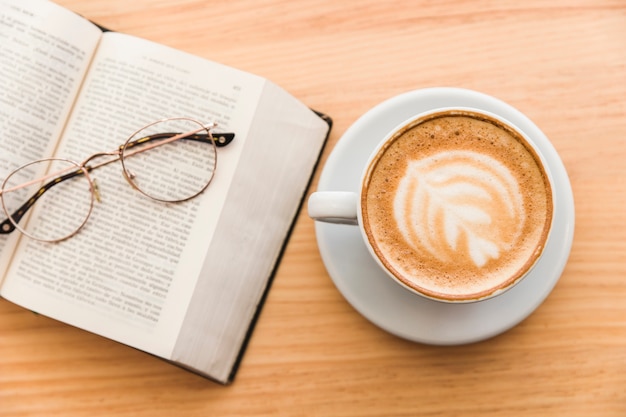 Xícara de café quente com arte de cappuccino latte e óculos sobre um livro aberto na mesa