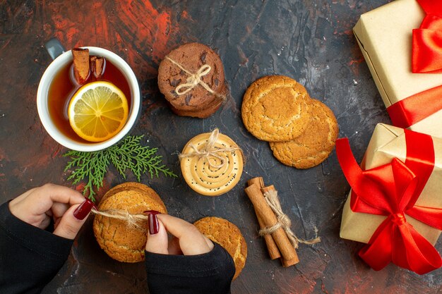 Xícara de chá de canela em pau de canela com biscoitos diferentes amarrados com corda em presentes de mão feminina na mesa vermelha escura