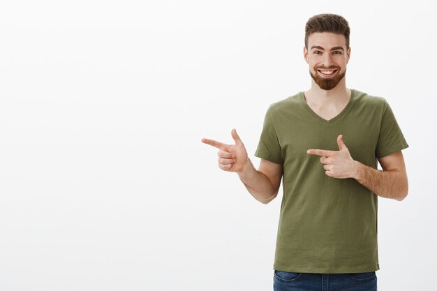 Yo olha isso se apresse. Retrato de um cara barbudo atraente entusiasmado e animado com uma camiseta verde-oliva sorrindo encantado ao apontar para a esquerda com pistolas de dedo para mostrar o produto incrível sobre a parede branca