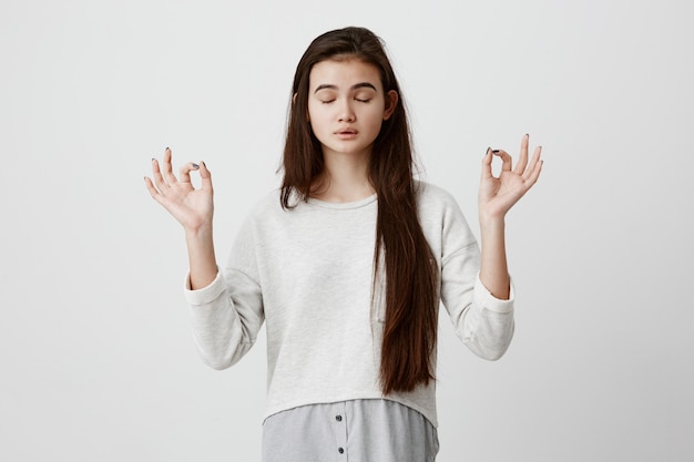 Foto grátis yoga e meditação. linda mulher morena vestida casualmente, mantendo os olhos fechados enquanto medita, sentindo-se relaxada, calma e pacífica