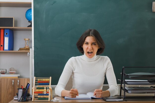 zangado olhando para a câmera jovem professora sentada na mesa com ferramentas escolares em sala de aula