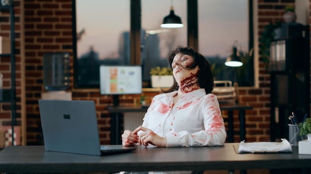 Foto grátis zumbi assustador sentado na mesa enquanto acenava para a videoconferência no laptop. monstro morto cerebral assustador com cicatrizes profundas e sangrentas no escritório tendo uma conversa online no computador moderno.