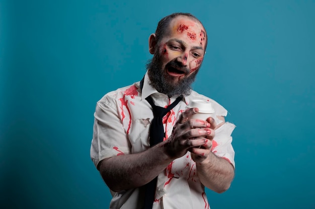 Foto grátis zumbi perigoso olhando para a xícara de café, querendo beber bebida e sendo agressivo no estúdio. monstro do mal assustador segurando o copo e tendo feridas sangrentas, diabo misterioso apocalíptico.