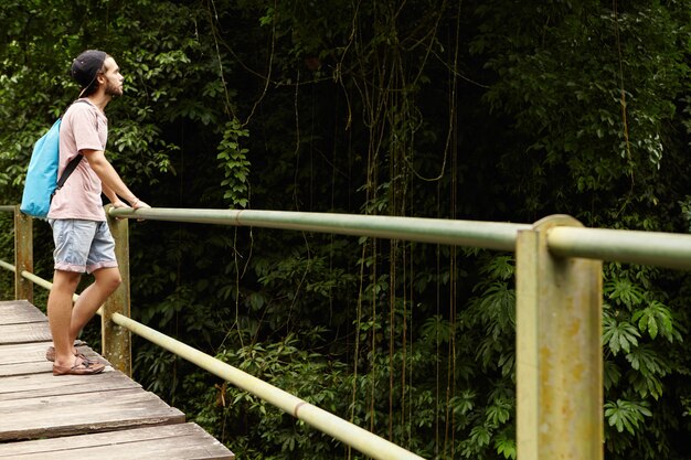 Abenteuer und Tourismus. Hübscher kaukasischer Student, der im Regenwald wandert. Junger Wanderer mit Rucksack, der auf Holzbrücke steht und grüne Wälder betrachtet