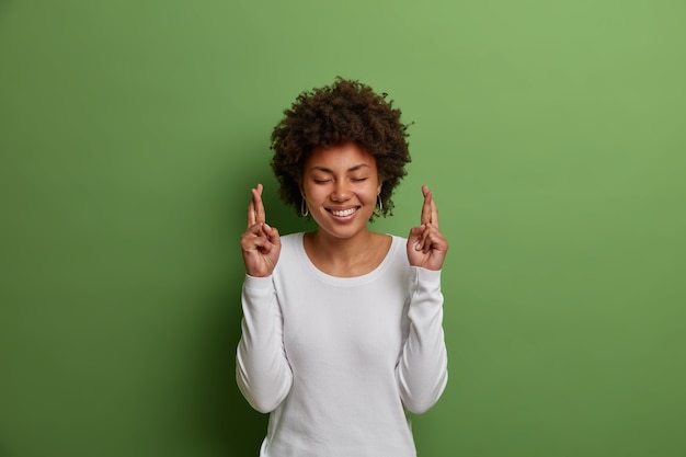 Abergläubische hoffnungsvolle frau mit afro-frisur, lächelt breit, drückt die daumen, damit träume wahr werden, macht wünsche und glaubt an ein besseres leben, trägt einen weißen pullover, isoliert an der grünen wand