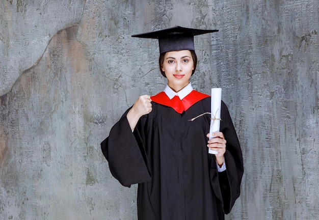 Abgeschlossene junge Frau im Kleid, das mit Universitätsdiplom aufwirft. Foto in hoher Qualität