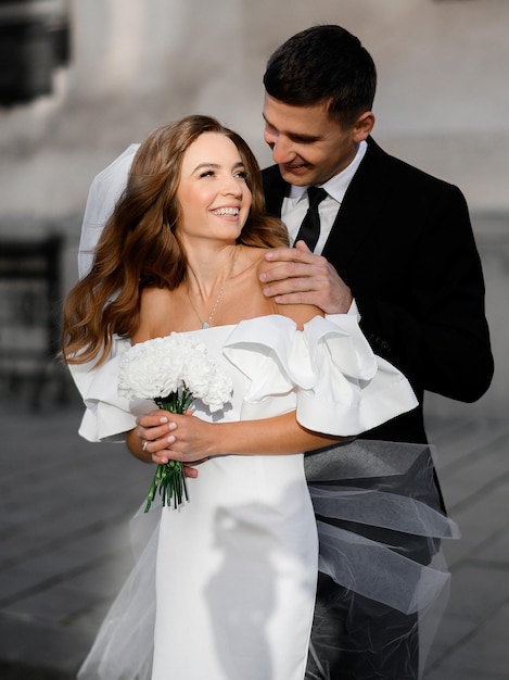 Abgeschnitten von einem hübschen Bräutigam im schwarzen Hochzeitsanzug, der die nackten Schultern einer glücklichen jungen Braut in Schleier und weißem Kleid berührt, die einen Blumenstrauß hält und sich auf der Straße in der Innenstadt ansieht