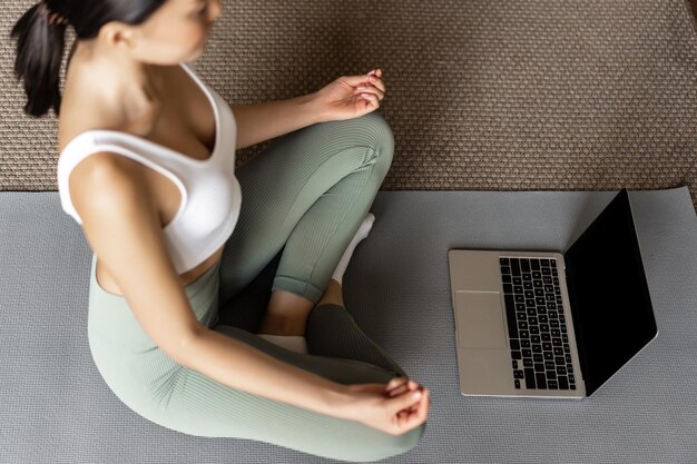 Abgeschnittene Aufnahme einer asiatischen Frau, die zu Hause auf einer Bodenmatte meditiert, Meditations-Podcast auf dem Laptop hört, Yoga-Online-Kurse praktiziert und Activewear trägt.