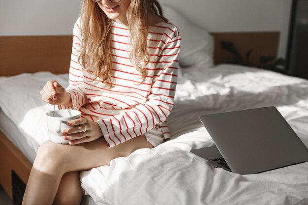 Abgeschnittene Aufnahme einer jungen Frau, die sich im Schlafzimmer entspannt, Tee trinkt und einen Laptop verwendet Lächelndes Mädchen, das mit einer Tasse im Bett sitzt