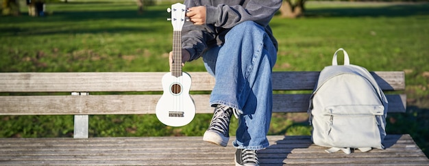 Kostenloses Foto abgeschnittene aufnahme eines jungen mädchens in turnschuhen und jeans, die ein ukulele-musikinstrument in der hand hält, während sie