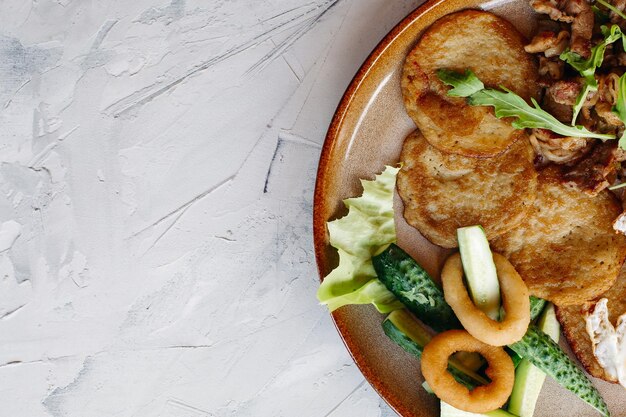 Abgeschnittene Draufsicht auf leckere Kartoffelpuncakes Frühstück mit Spiegeleiern Frische grüne Gurken, gebratene goldene Zwiebeln und knuspriges Fleisch, serviert mit köstlichen Salatblättern, die auf brauner Tonplatte luieren