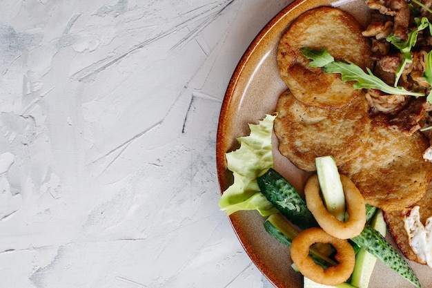 Abgeschnittene Draufsicht auf leckere Kartoffelpuncakes Frühstück mit Spiegeleiern Frische grüne Gurken, gebratene goldene Zwiebeln und knuspriges Fleisch, serviert mit köstlichen Salatblättern, die auf brauner Tonplatte luieren